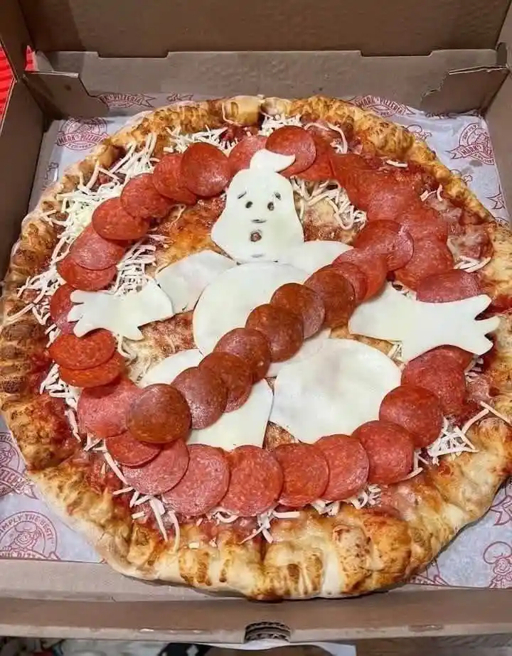 A pepperoni pizza in a cardboard box is decorated with cheese slices shaped like a ghost and pepperoni arranged in a circle…