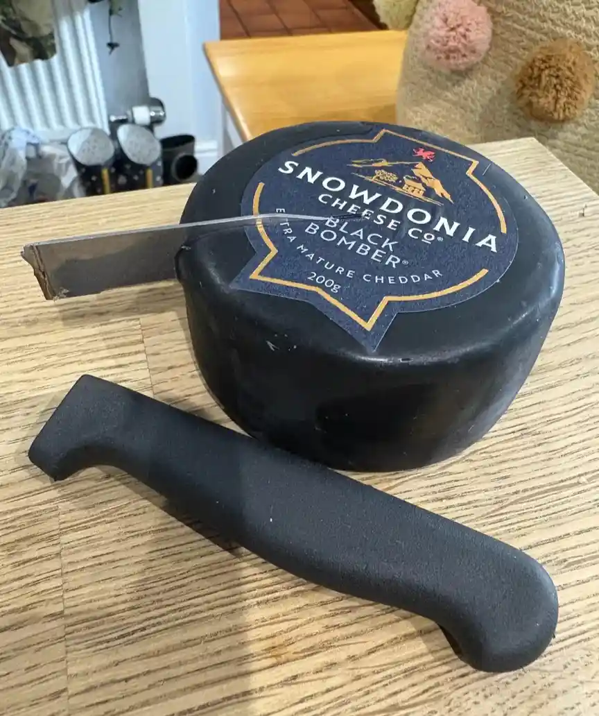 A round, black-wrapped cheese labeled 'Snowdonia Cheese Co. Black Bomber Extra Mature Cheddar 200g' sits on a wooden table…