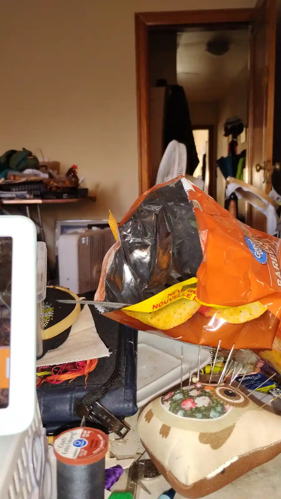 A messy room with craft supplies like thread, a pin cushion, and an embroidery hoop. A large orange bag of tortilla chips is…