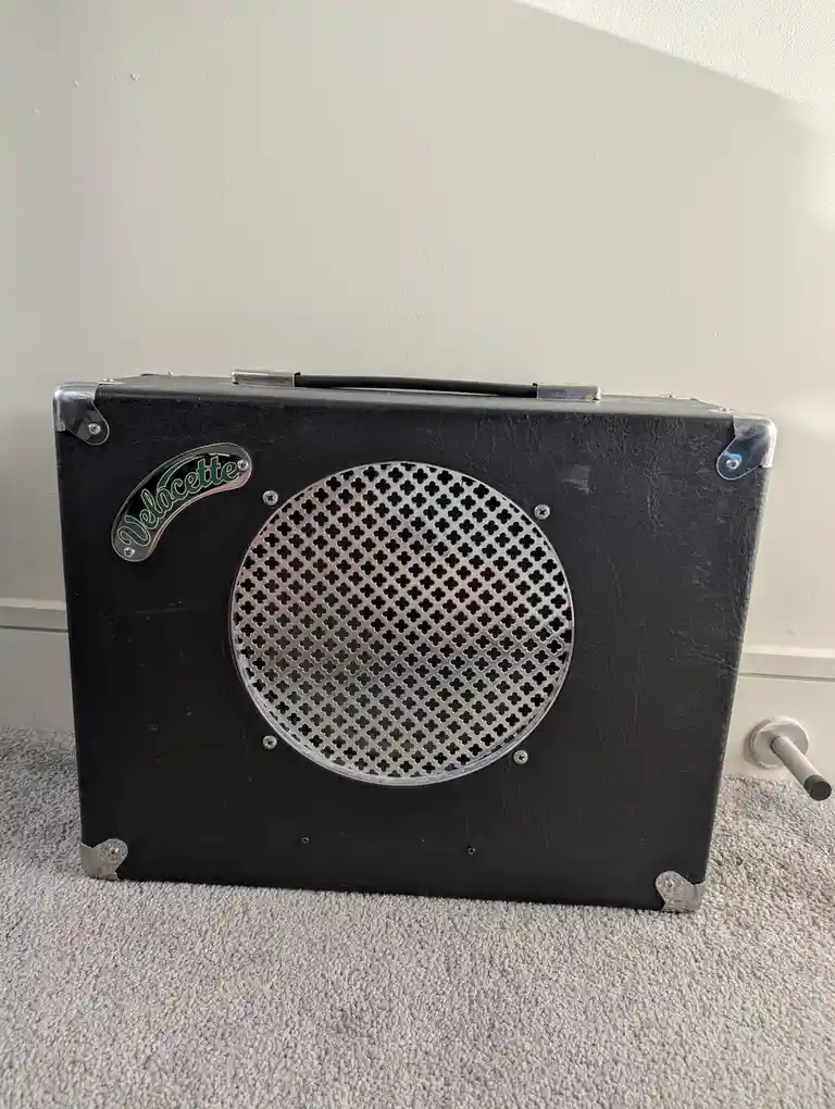 A black, suitcase-style guitar amplifier with a circular metal grille featuring a crosshatch pattern and a 'Velocette' logo…