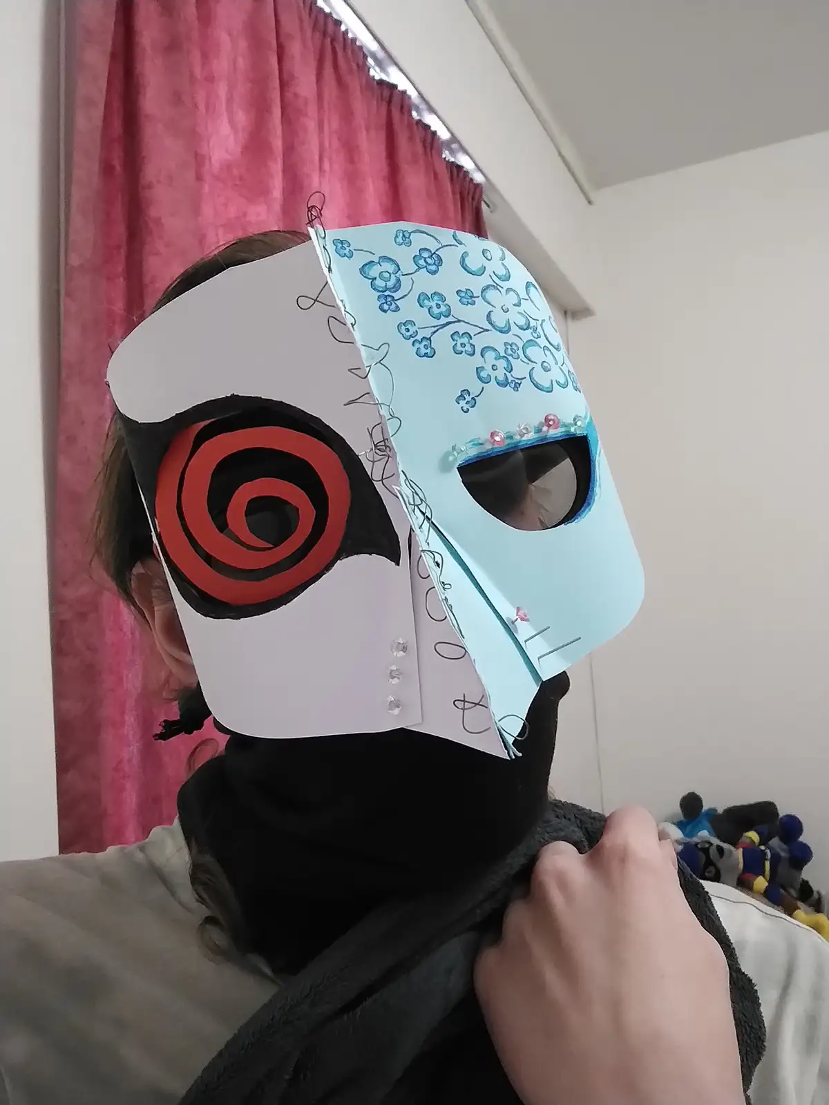 A person wears two handmade paper masks over their face, one half white with a red spiral eye and the other half light blue…