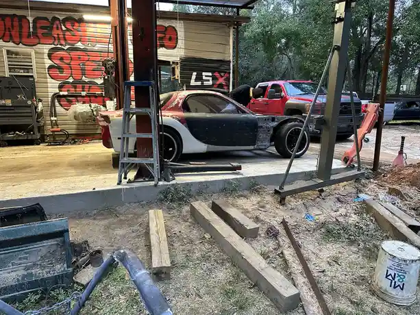A partially disassembled red and white race car sits on a concrete platform in a cluttered garage, surrounded by tools, wood…