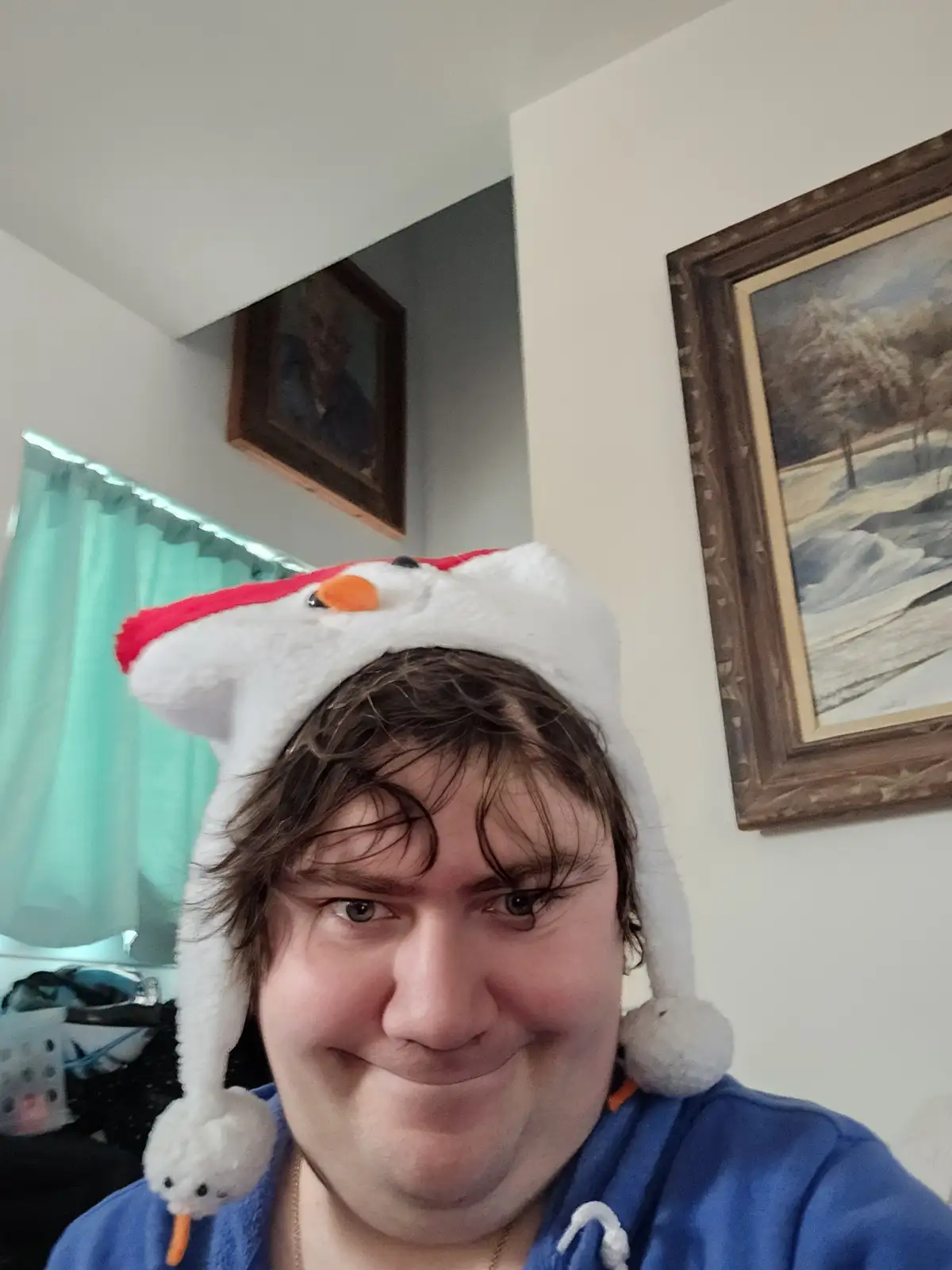 A person with messy brown hair and a blue hoodie wears a white, fluffy snowman hat with a red rim, black eyes, and an orange…