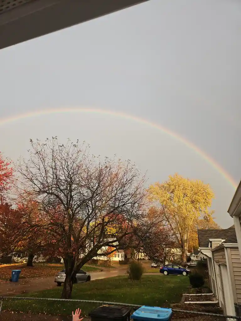 A person's hand is visible at the bottom of the frame, reaching up toward a full, vivid rainbow arching across a gray sky…