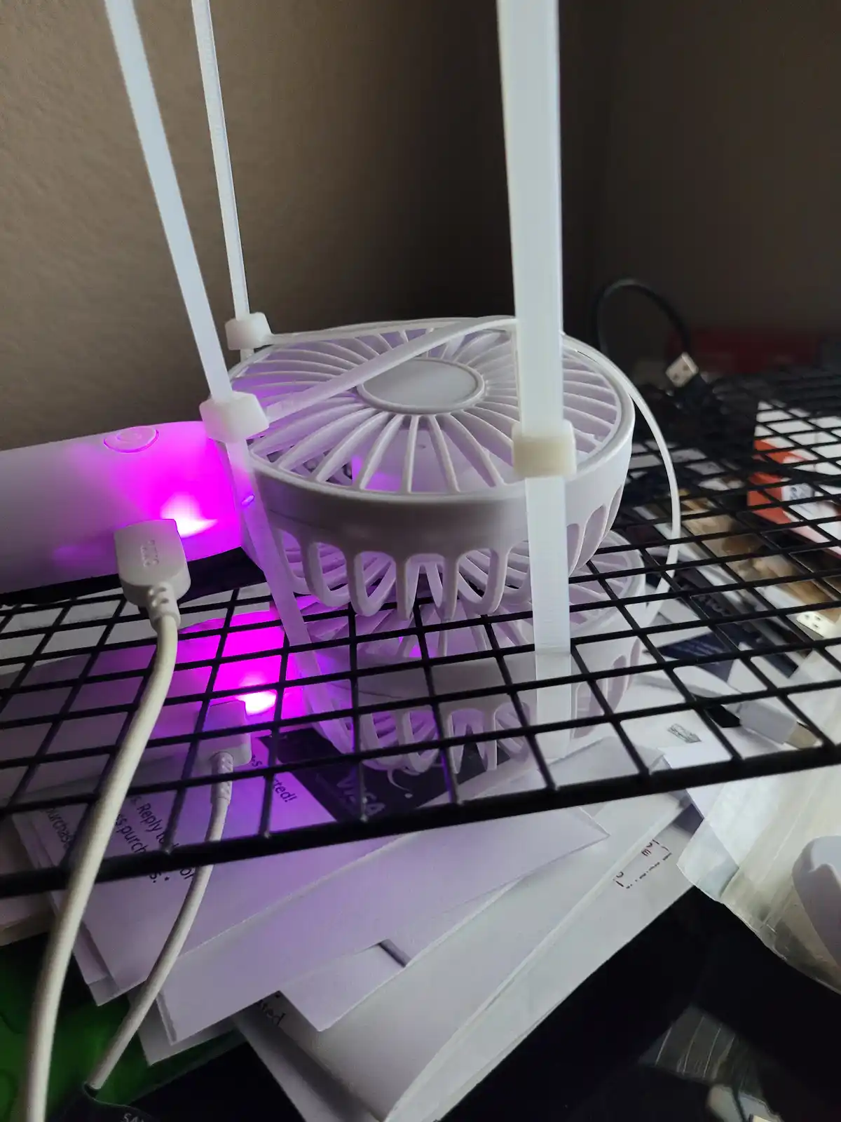 A white USB-powered desk fan is mounted vertically using white zip ties on a black wire shelf, with a purple LED light…