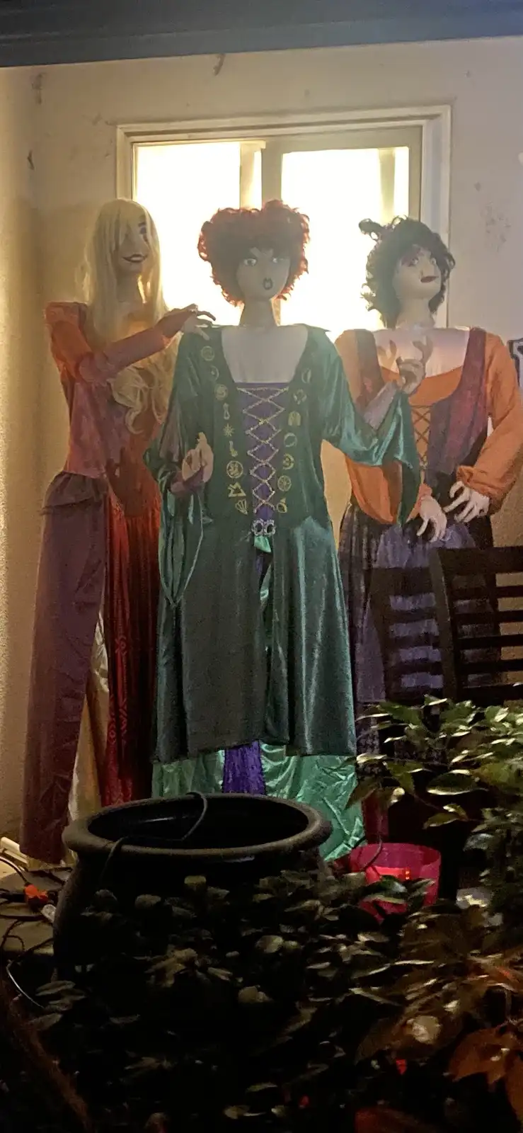 Three plastic witch figurines in Halloween costumes stand indoors near a window, with a cauldron in front. The central witch…