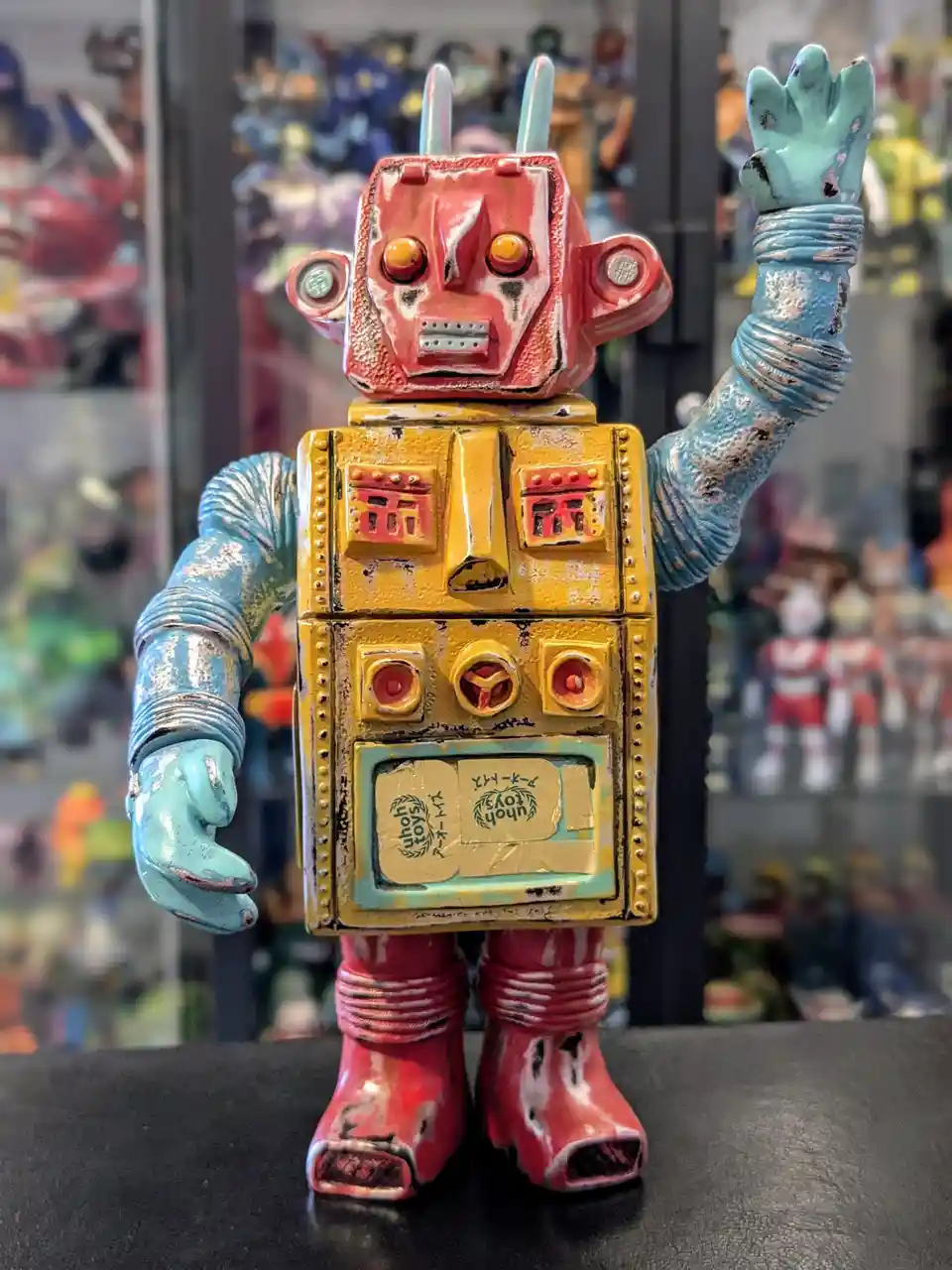 A vintage-style, weathered robot toy with a red head, yellow torso, and blue arms stands on a black surface, one arm raised…