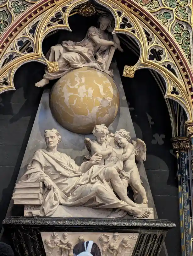 A close-up of an ornate, gilded Gothic-style tomb or memorial featuring a reclining marble statue of a man with books, two…