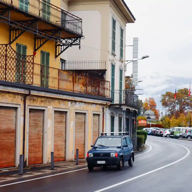 A blue Fiat Panda drives down a curving European street past old buildings with green shutters and rusted balconies. The…