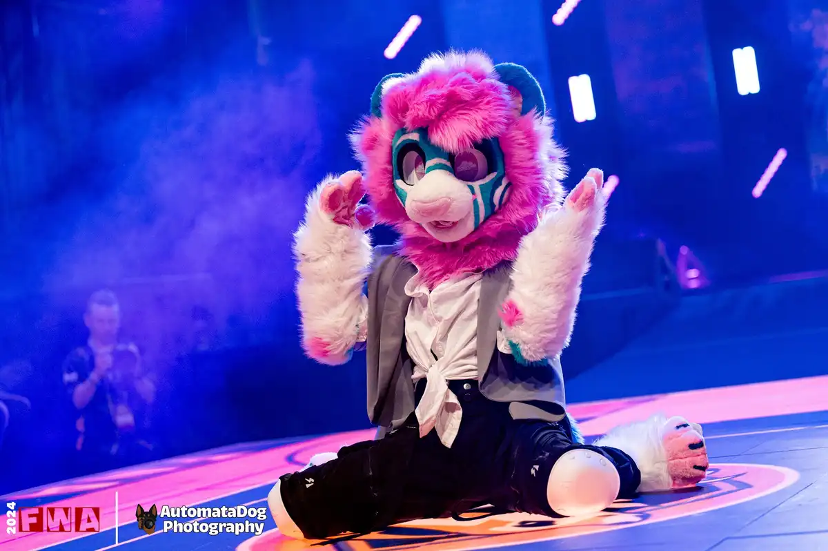 A person in a pink and teal fursuit with large eyes and white paws is sitting on a stage, hands raised in a gesture of…