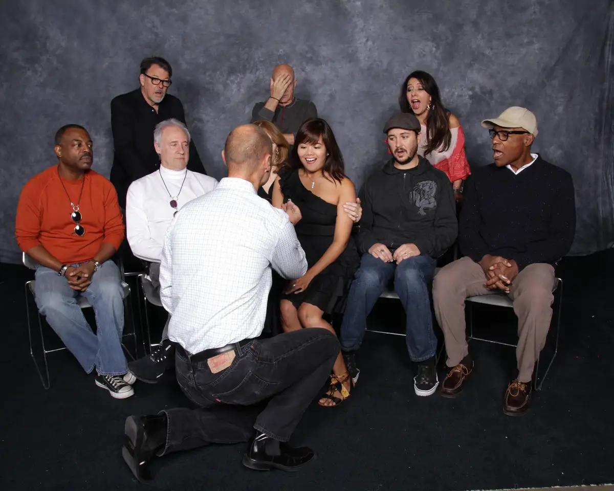 A man kneels before a woman seated among a group of people, all reacting with varied expressions of shock, amusement, or…