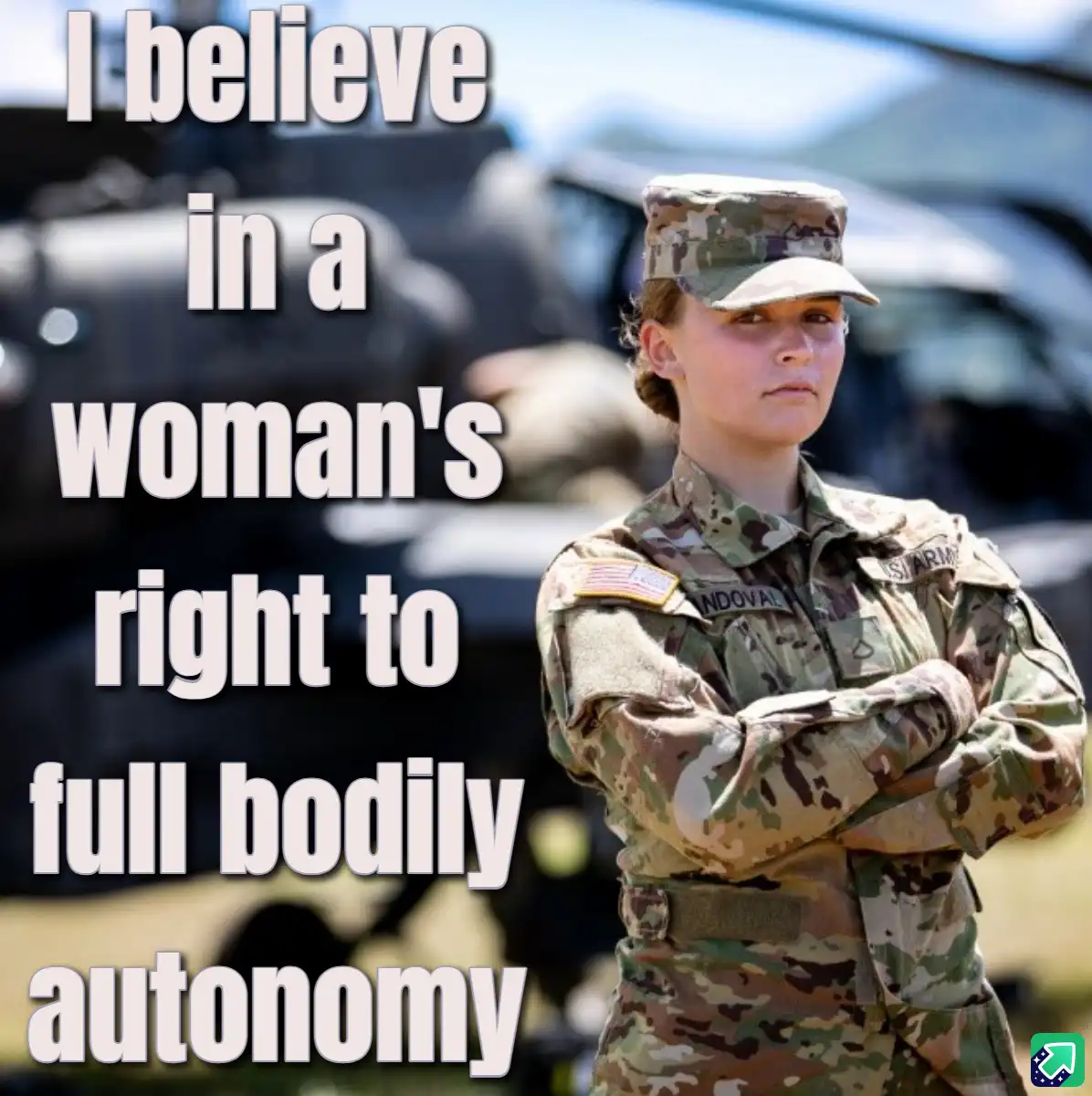 A woman in U.S. Army camouflage uniform stands with arms crossed, looking serious, in front of blurred military vehicles…