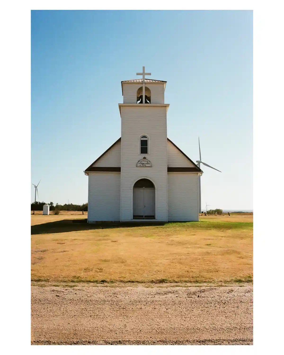 A white wooden church with a bell tower and cross stands in a dry, open field under a clear blue sky, with several wind…