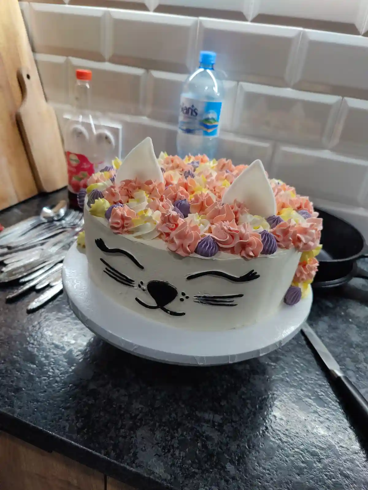 A round white-frosted cake sits on a kitchen counter, decorated with black icing to resemble a cat's face, complete with…