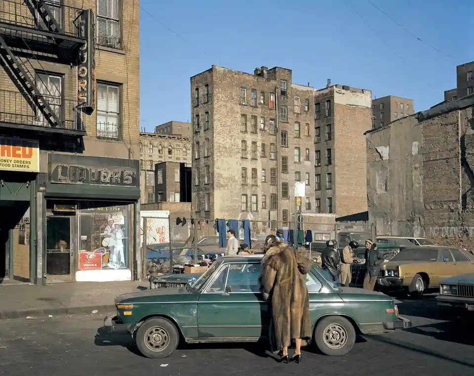 A vintage street photograph shows a woman in a long fur coat leaning against a green car parked on a gritty urban street…