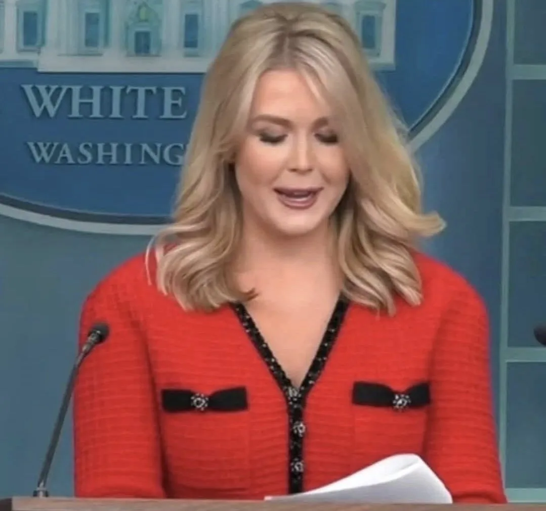 a woman with blonde hair wearing a red jacket with black trim and bows stands at a podium in front of a blue backdrop that…