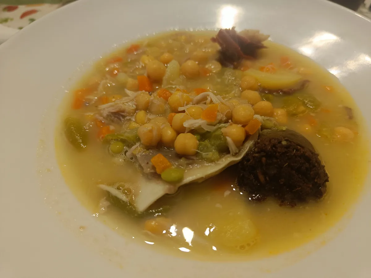 a close-up photo of a white bowl filled with a yellowish broth containing chickpeas, diced carrots, shredded meat, green…