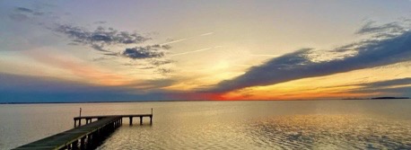 A serene sunset over a calm body of water with a long wooden pier extending into the distance. The sky is painted with warm…