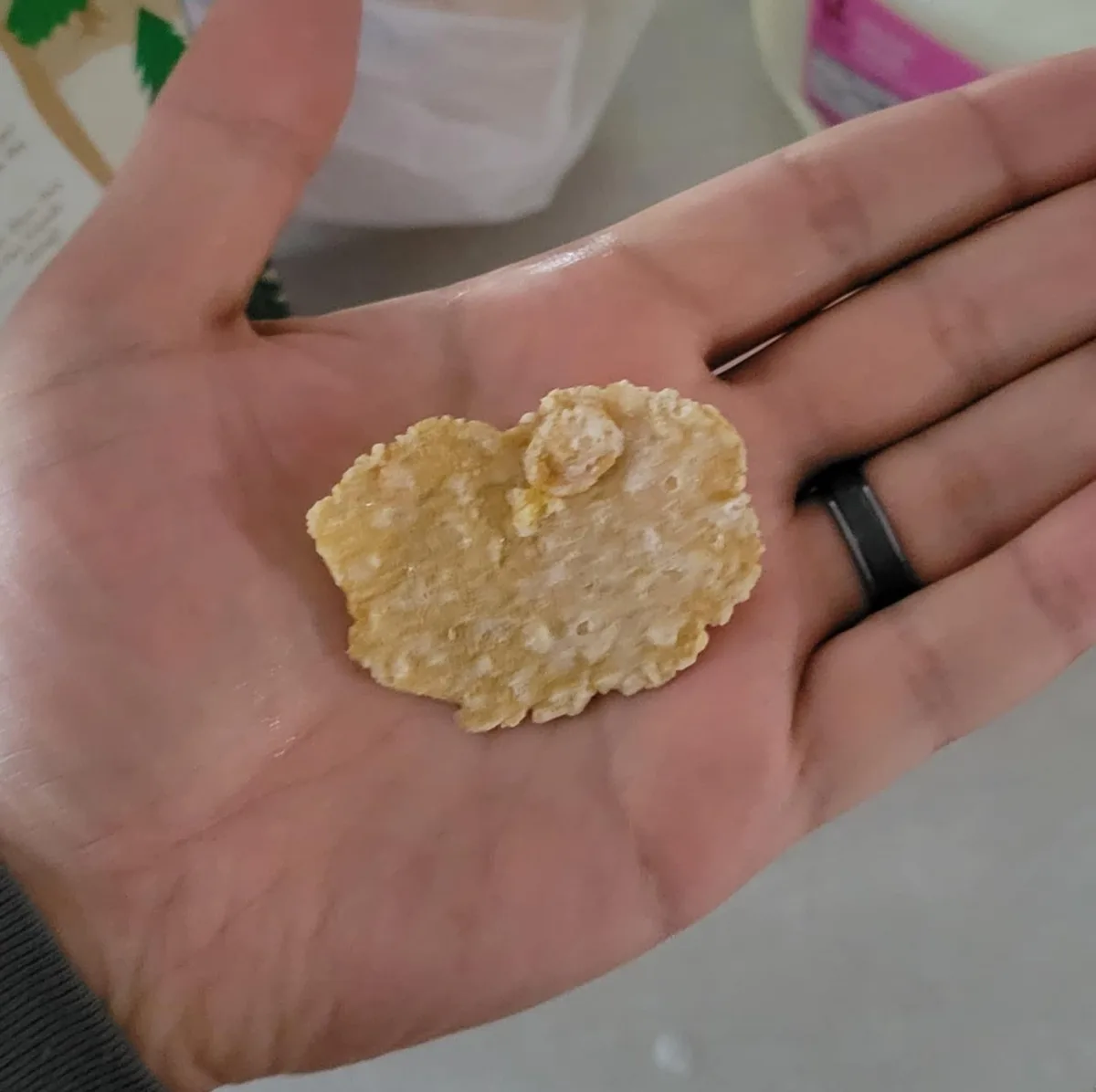 A person's open palm holds a single, irregularly shaped heart-shaped cereal piece, which appears slightly broken or…