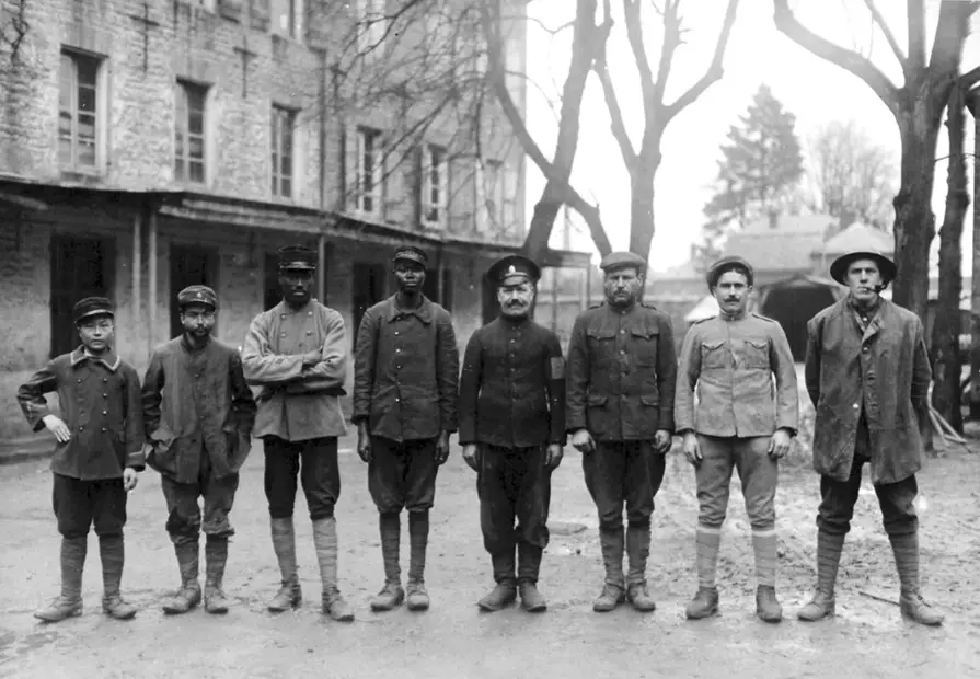 A black-and-white historical photo shows eight men in early 20th-century military uniforms standing in a line outdoors. They…