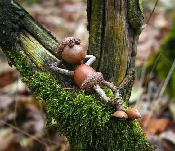 A whimsical, handcrafted figurine made from acorns and twigs is lounging on a moss-covered tree branch, appearing to rest…
