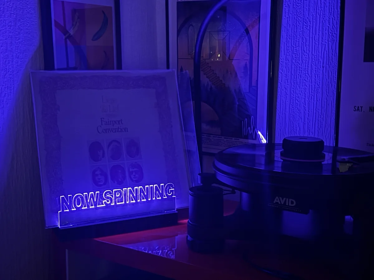 A vinyl record player sits under blue light next to a 'NOW SPINNING' neon sign, with a Fairport Convention album cover…