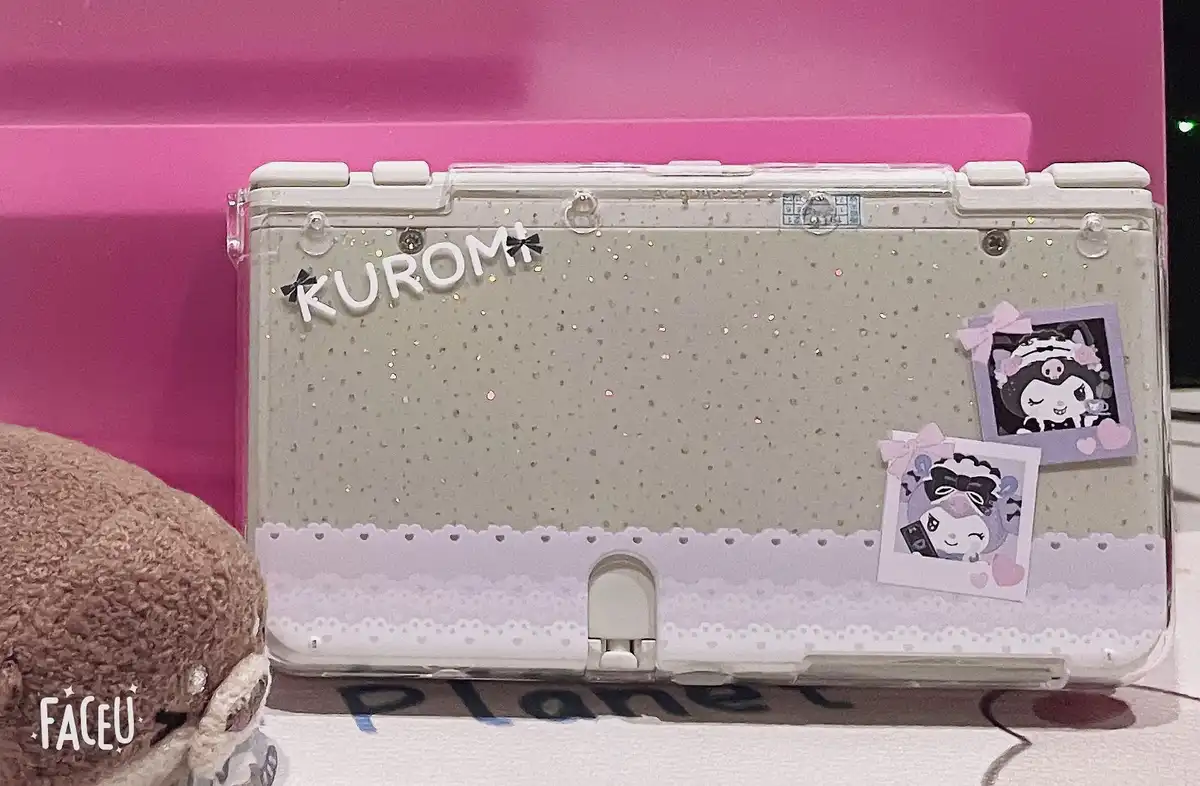 A decorated Nintendo 3DS console with a clear case, glitter, and two Kuromi character stickers, with the word 'KUROMI'…