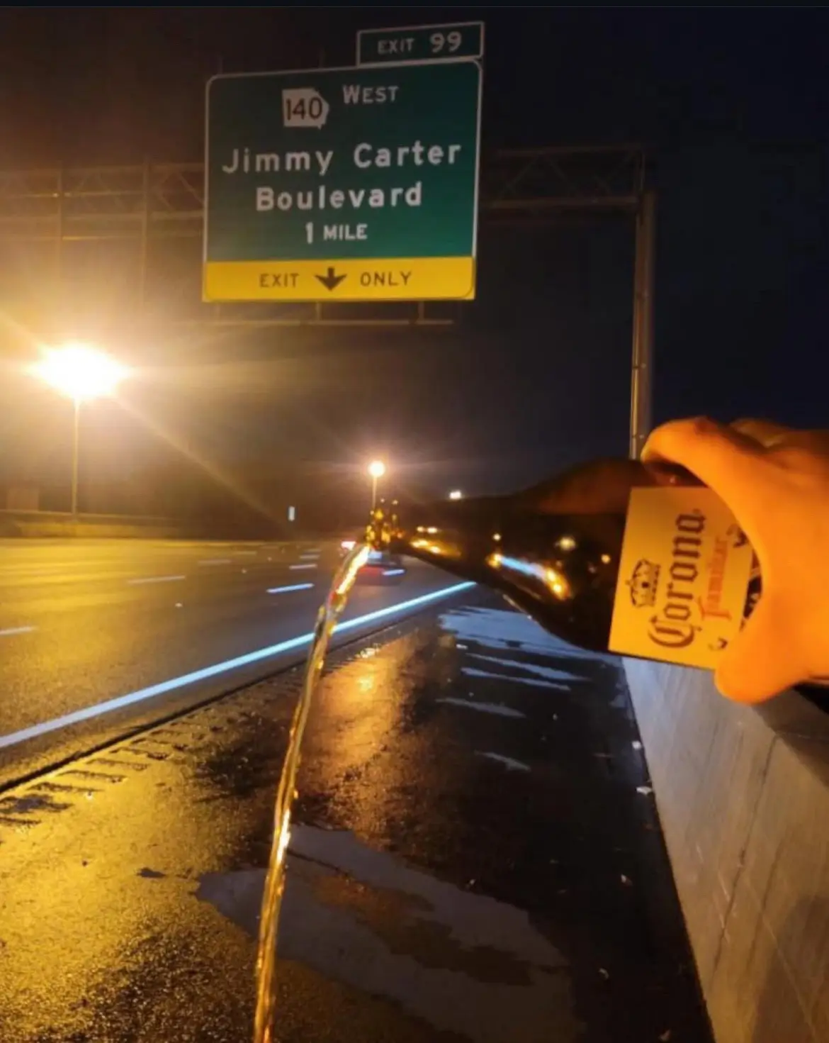 A hand pours a bottle of Corona beer onto a wet highway shoulder at night, with a green highway sign for 'Jimmy Carter…