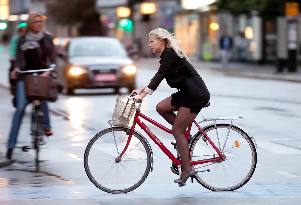 A blonde woman in a black business suit and high heels rides a red bicycle with a basket on a wet city street, looking…