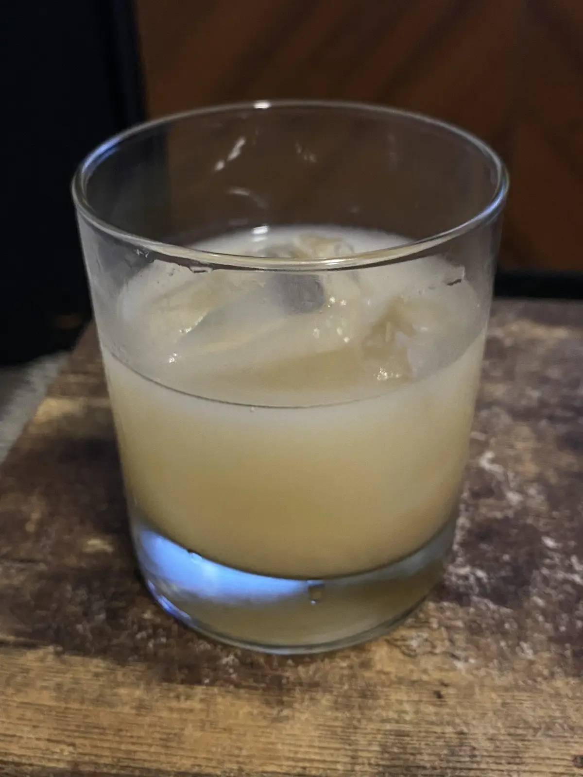 A close-up photo shows a rocks glass filled with a pale yellow liquid and ice cubes, sitting on a wooden surface. The…