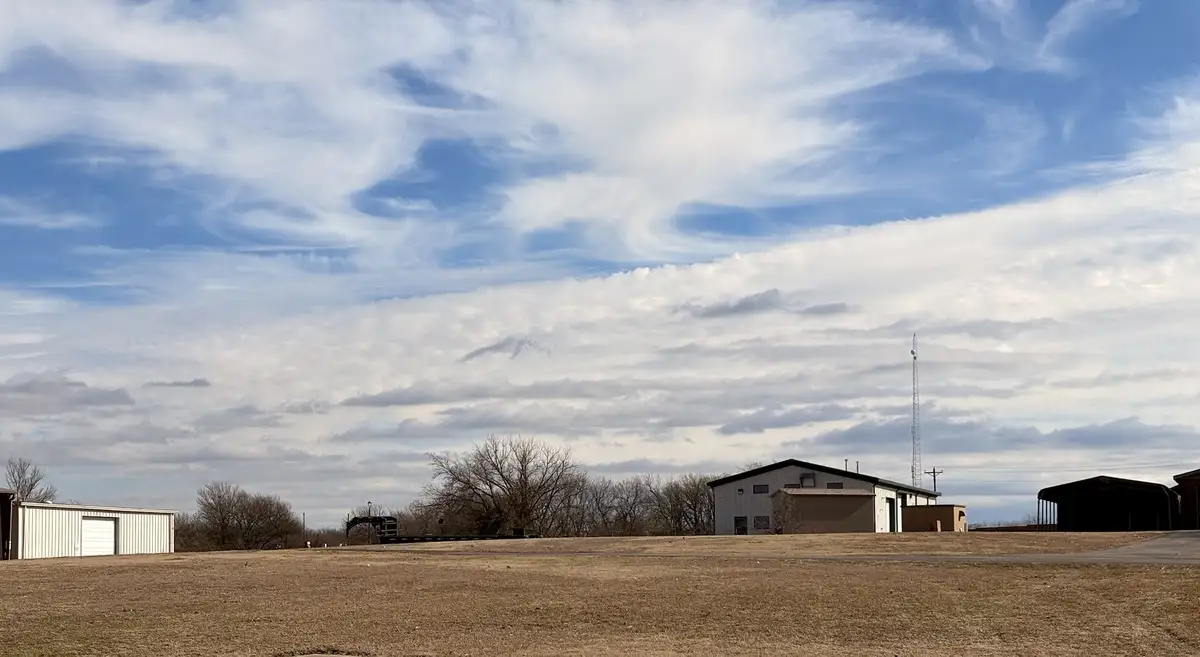 A wide shot of a rural landscape with several metal buildings, bare trees, and a vast sky filled with wispy clouds that form…