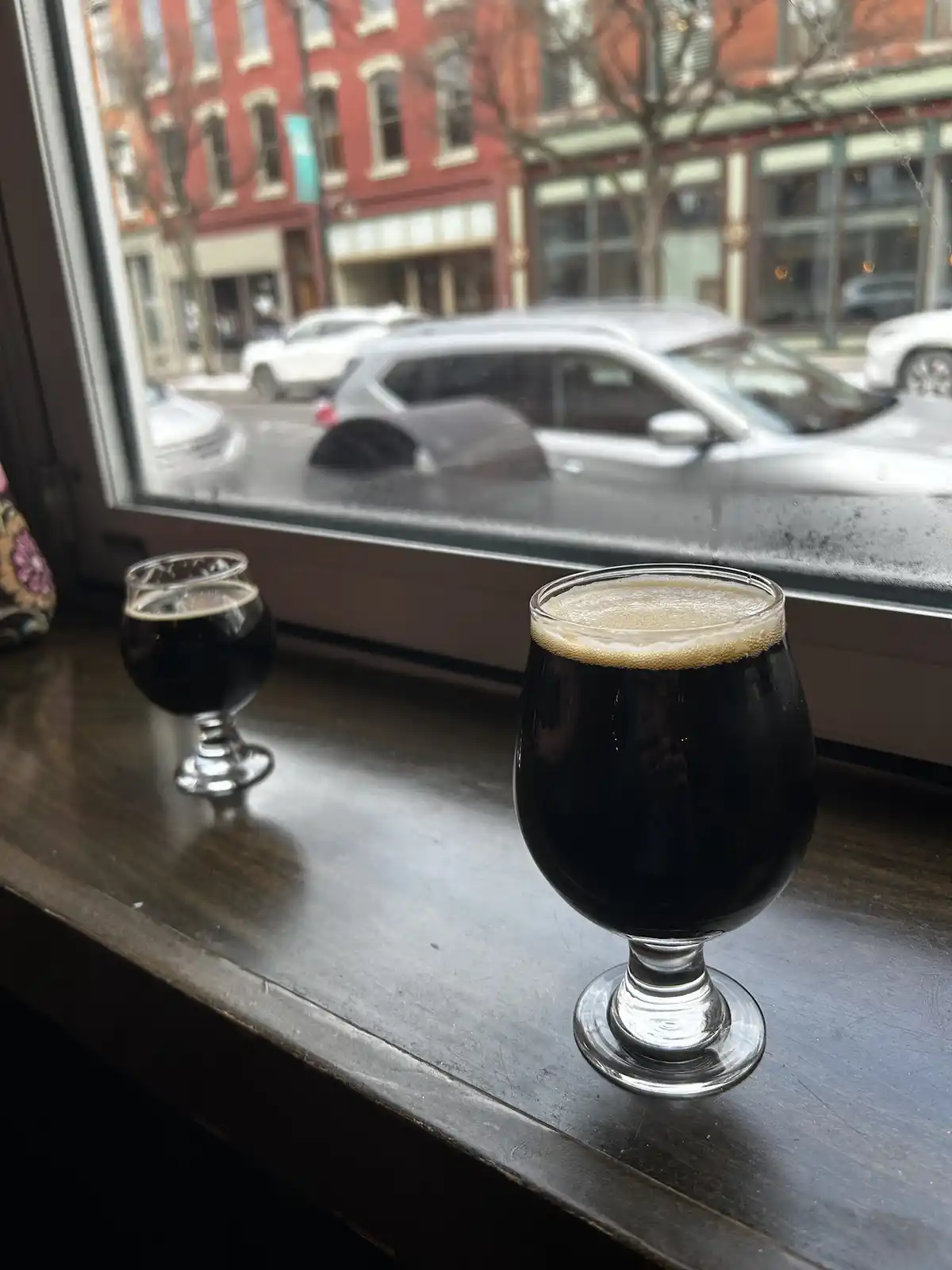 Two dark beers in stemmed glasses sit on a wooden windowsill, with a rainy street and red brick buildings visible through…