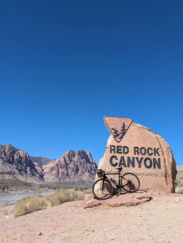 A black road bicycle is leaning against a large, reddish-brown rock sign that reads 'RED ROCK CANYON National Conservation…