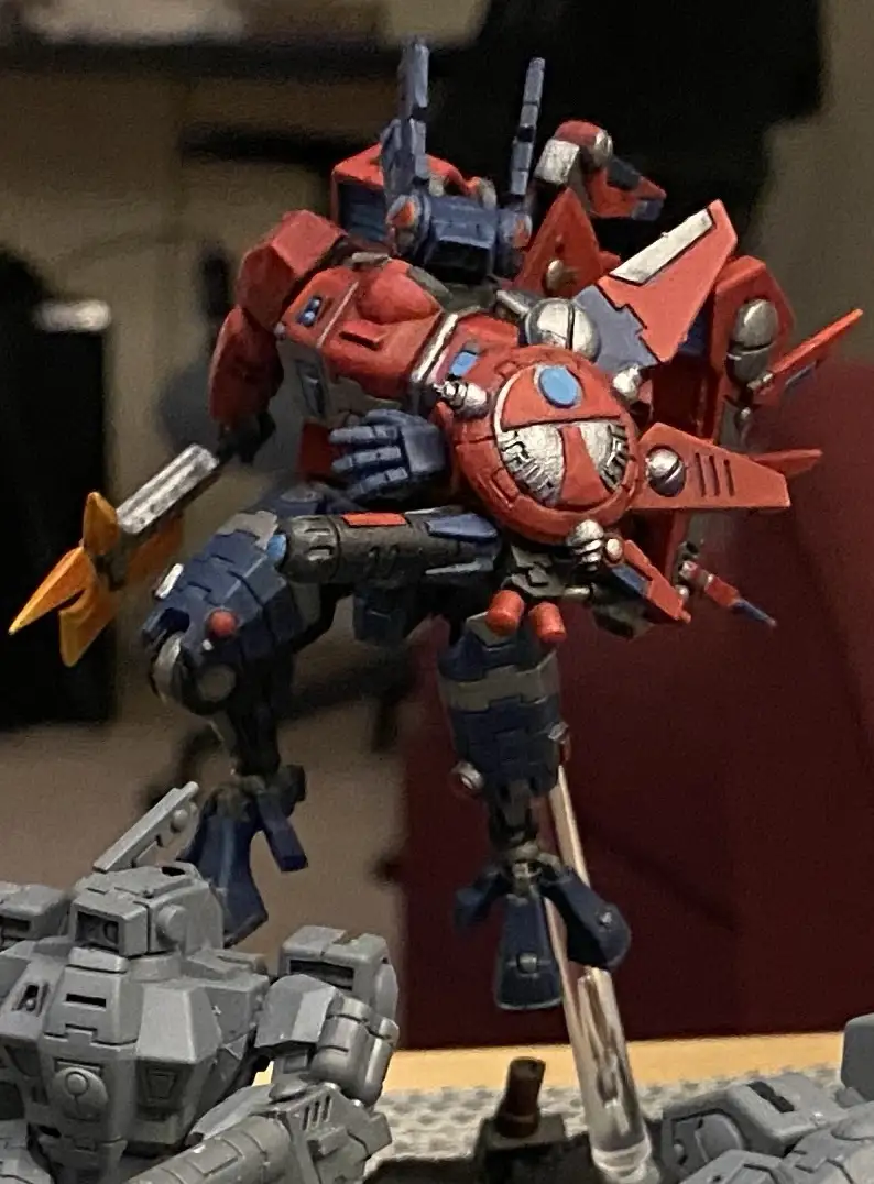 A red and blue painted transformer action figure stands in a dynamic pose, holding a weapon, with other grey robot figures…