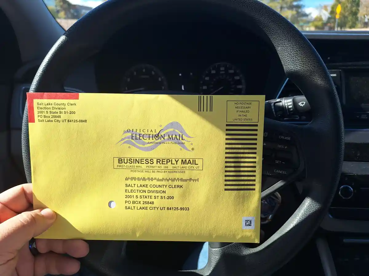 A hand holds a yellow Salt Lake County Clerk Election Division official election mail business reply envelope in front of a…
