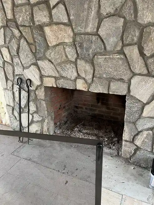 A photo of an empty stone fireplace with a metal screen door in front, brick interior, and fireplace tools leaning against…