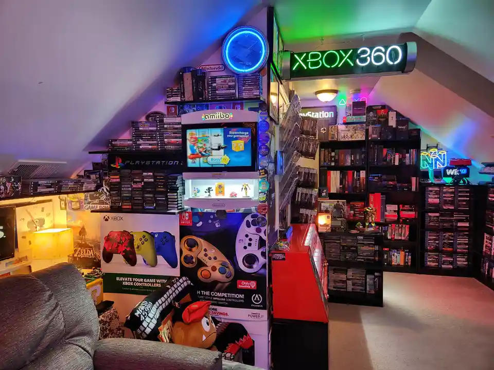 A brightly lit, cluttered gaming room filled with shelves of video game consoles, controllers, and collectibles, featuring a…