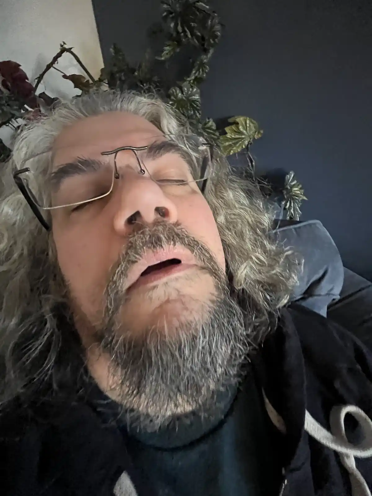 A man with long gray hair and a beard, wearing glasses, lies on his back with eyes closed and mouth slightly open, appearing…