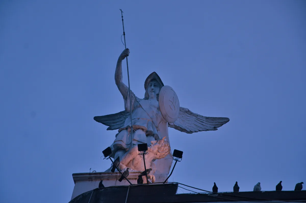 A low-angle shot of a large, white stone statue of an angel with wings, holding a spear and shield, set against a dusky blue…