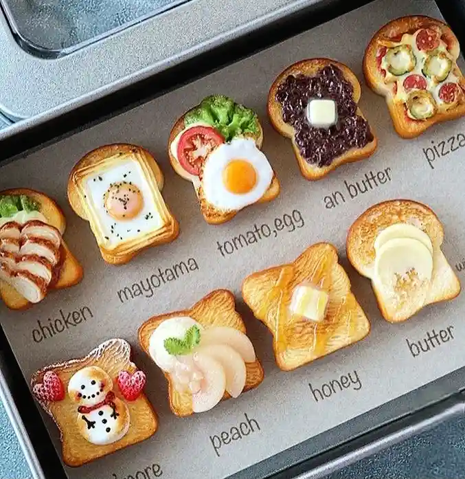 An overhead shot of a baking tray holding miniature, intricately decorated bread slices resembling various breakfast foods…