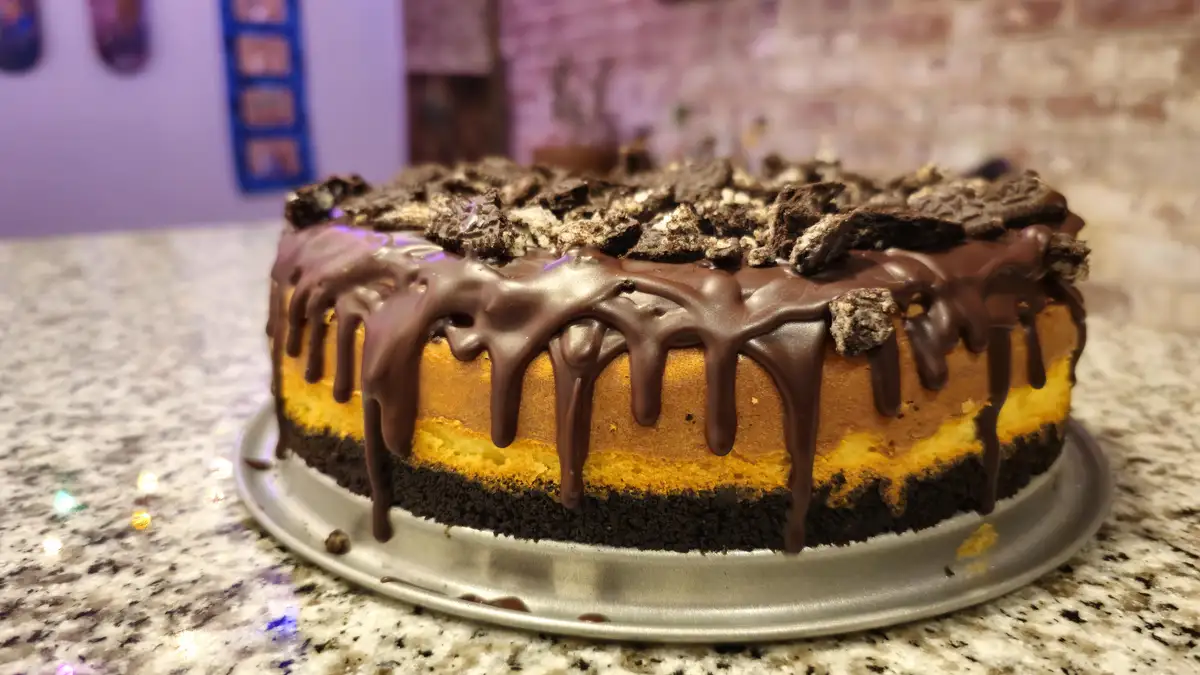 a close-up photo of a round cheesecake with a dark chocolate ganache drip cascading down its sides and crushed chocolate…