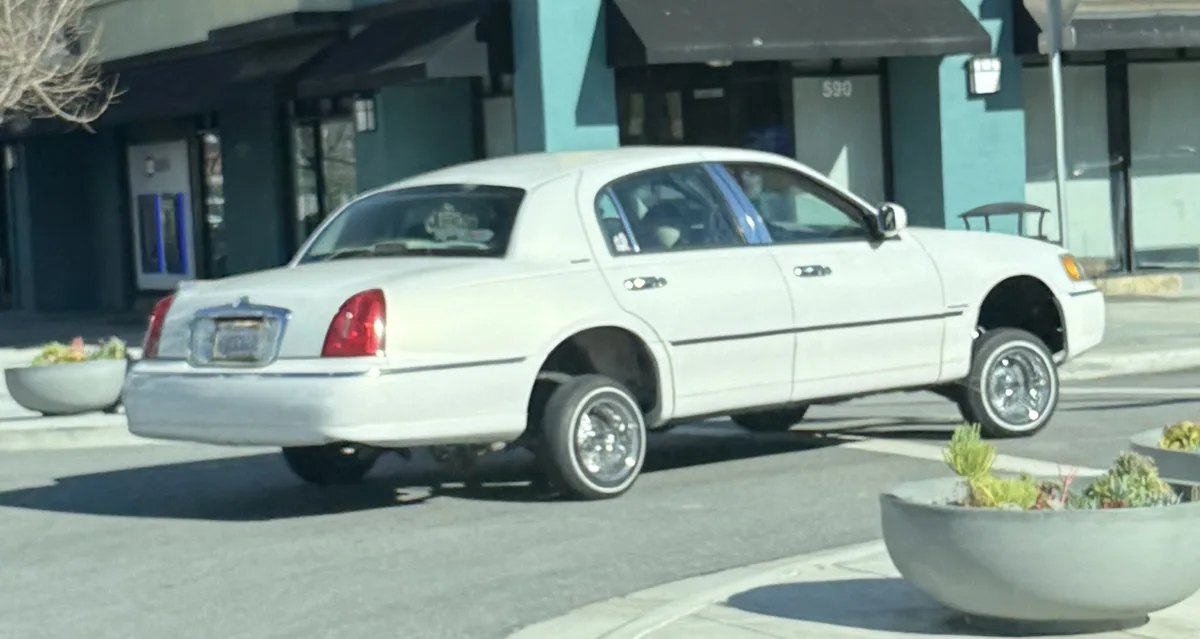 A white sedan is parked on a street in front of a building with the number 590. The car's rear wheels are missing, and the…