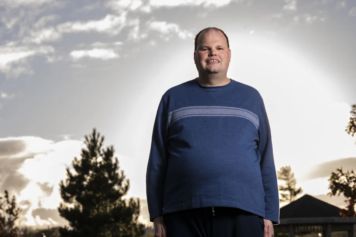 A man in a blue long-sleeved shirt stands outdoors against a cloudy sky with trees and a structure in the background. He has…
