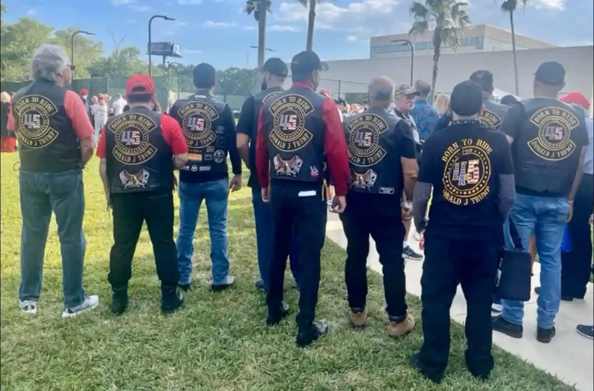 A group of men stand with their backs to the camera, wearing black vests with a large circular patch that reads 'BORN TO…