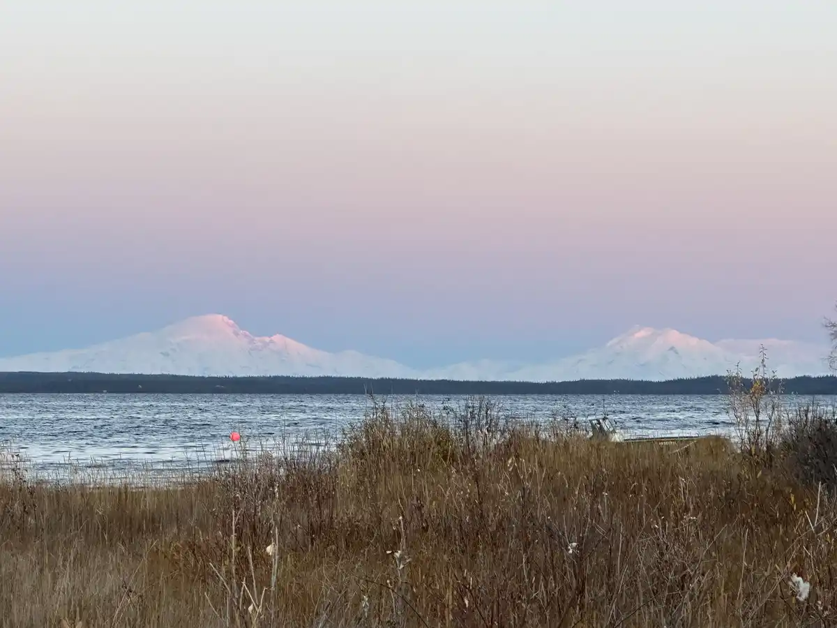 A serene landscape photo shows snow-capped mountains under a soft pink and blue twilight sky, with a calm lake in the middle…