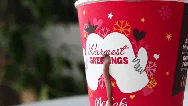A red McDonald's McCafé holiday cup with a white mitten graphic and "Warmest GREETINGS" text has a brown, viscous substance…