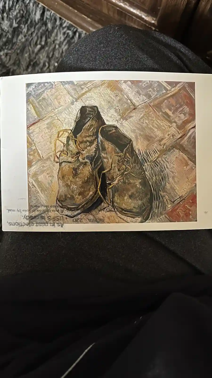 A printed reproduction of Vincent van Gogh's painting 'A Pair of Shoes' is shown resting on a dark gray fabric surface. The…