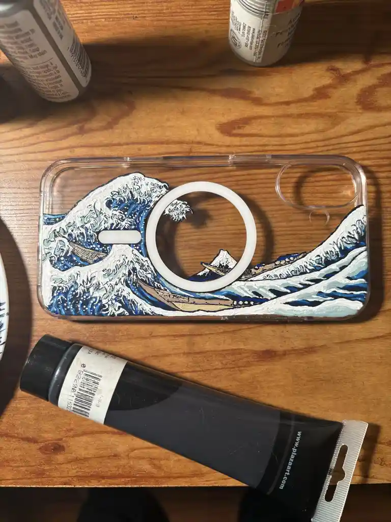 A clear phone case with a hand-painted rendition of Katsushika Hokusai's 'The Great Wave off Kanagawa' is shown on a wooden…