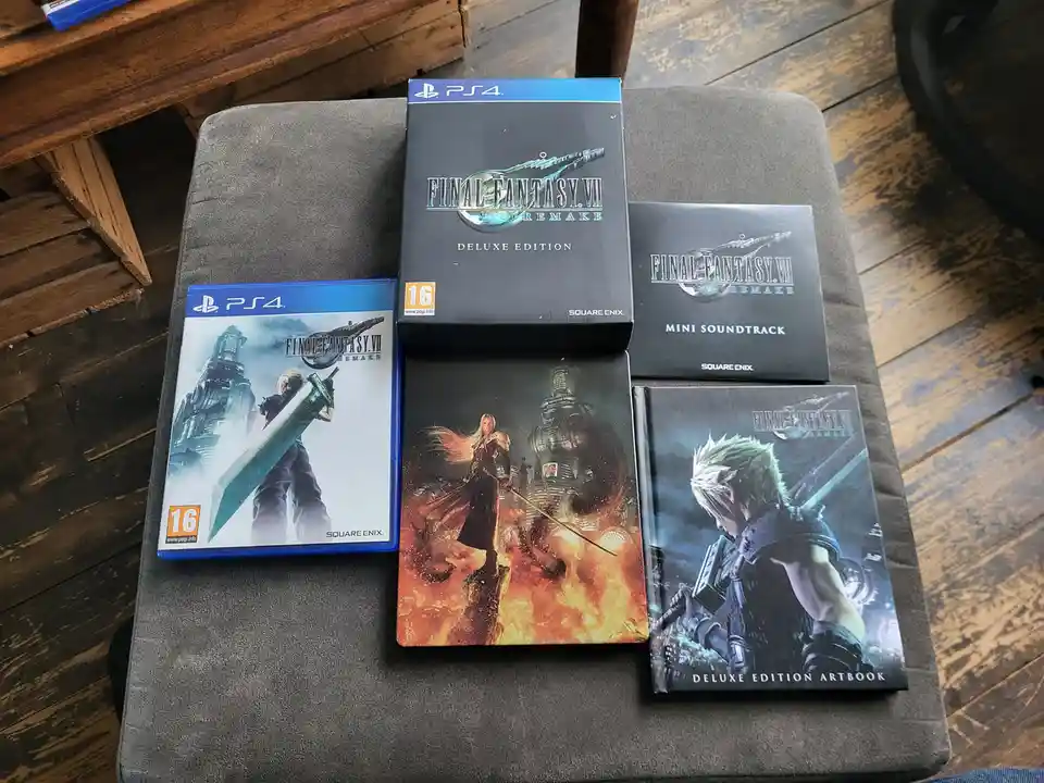Five physical items from the Final Fantasy VII Remake for PS4 are arranged on a grey cushion: a standard game case, a black…