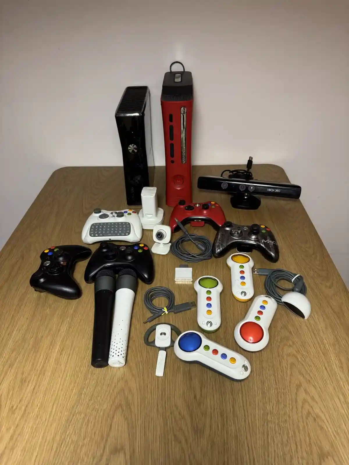A wooden table displays a large collection of Xbox 360 hardware, including two consoles (black and red), multiple…